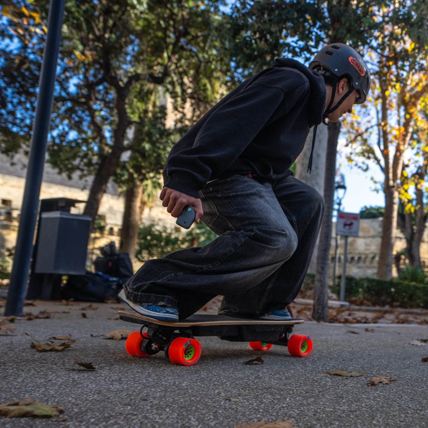 Person riding Meepo Edge electric skateboard with orange wheels on a leaf-covered path